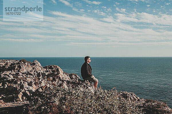 Junger Mann schaut von einer Klippe in Portugal im Sommer auf das Meer