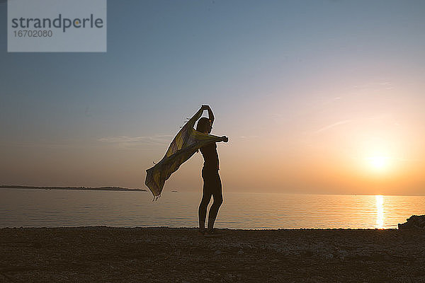 Silhouette einer Frau  die mit ihrem Schal am Strand steht