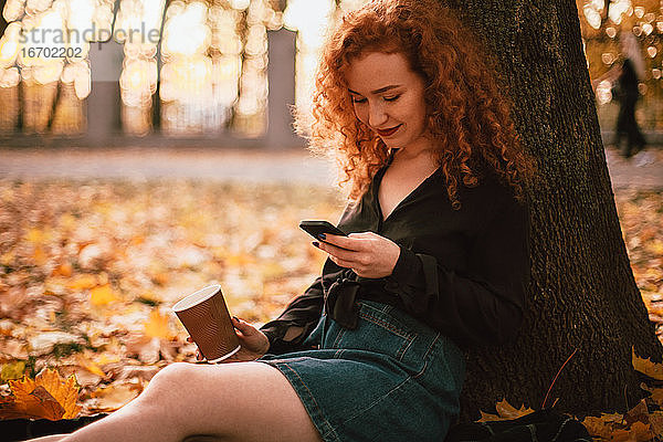 Glückliche junge Frau  die ihr Smartphone benutzt  sitzt im Herbst an einem Baum im Park