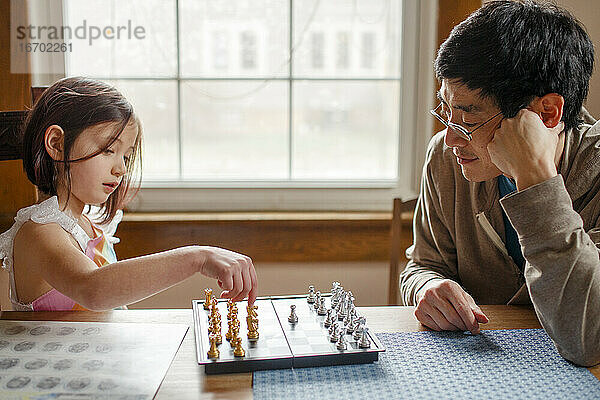Ein kleines Kind spielt Schach mit lächelndem Vater im Fensterlicht