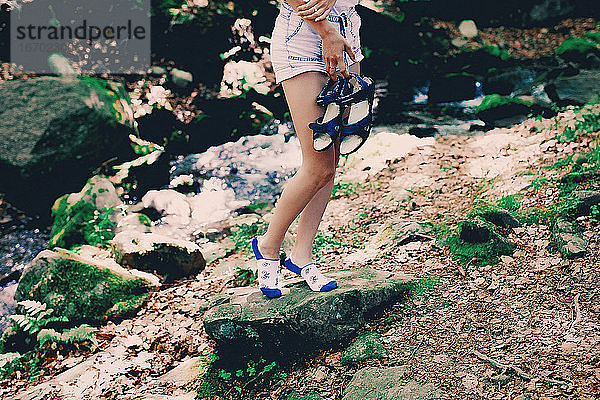 junges Hipster-Mädchen in bedruckten hellen Socken  das auf einem Felsen am Fluss im Wald sitzt