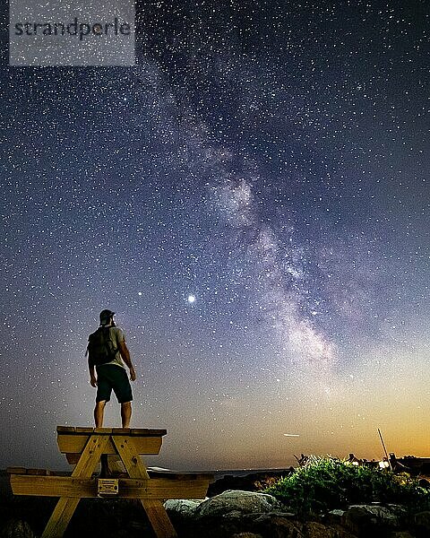 Silhouette eines Mannes  der auf einem Picknicktisch unter der Milchstraße die Sterne betrachtet.