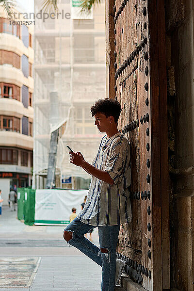 Der junge Afro lehnt an einem Holztor und schaut auf sein Handy