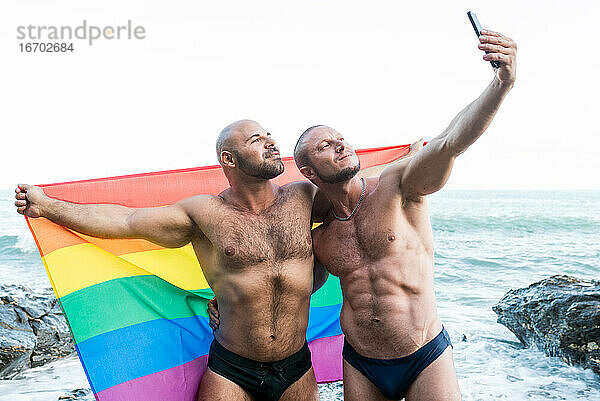 Lgbt Stolz Konzept. Homosexuell Bär Paar hält Regenbogenflagge mit offenen Armen ein Selfie-Foto zu nehmen.