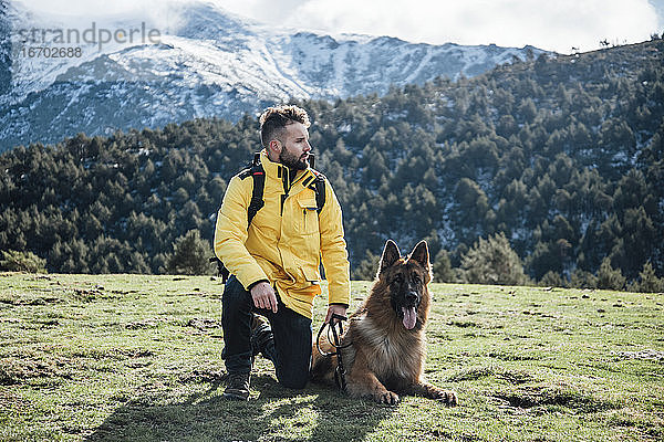 Junger Mann mit gelber Jacke und Rucksack spielt mit deutschem Schäferhund in den Bergen.