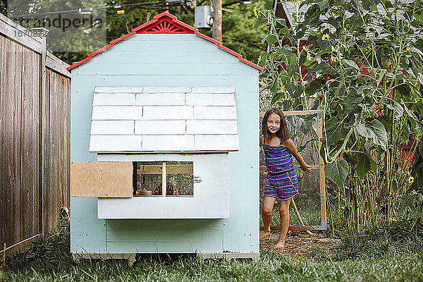 Ein stolzes kleines Mädchen steht vor der offenen Tür eines Hühnerstalls im Garten