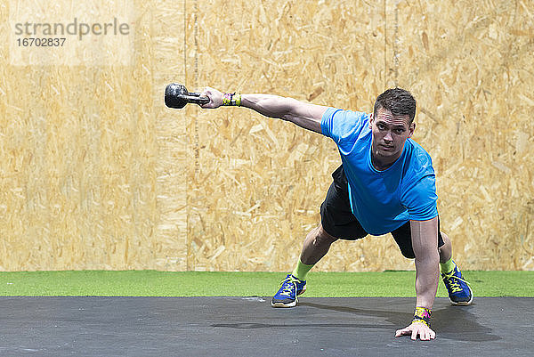 Vorderansicht eines sportlichen kaukasischen Mannes in Sportkleidung  der in einem Fitnessstudio Cross-Training betreibt  Krafttraining mit Kettlebells  der sich auf einen Arm stützt und mit dem anderen Arm Kettlebells hebt