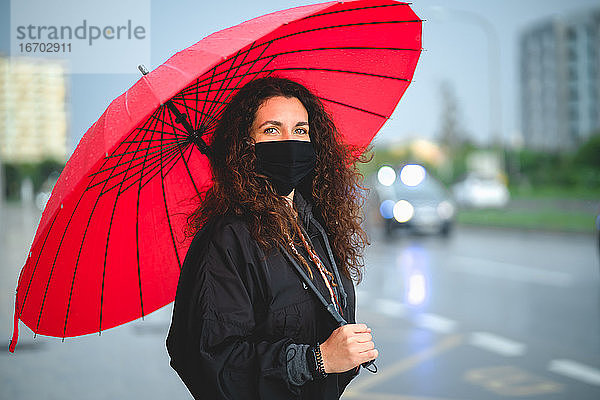 Porträt einer Frau mit einer Maske und einem roten Regenschirm  die in die Kamera schaut