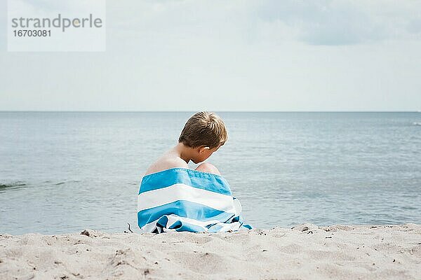 Ein kleiner Junge saß in ein gestreiftes Handtuch gewickelt allein am Strand.
