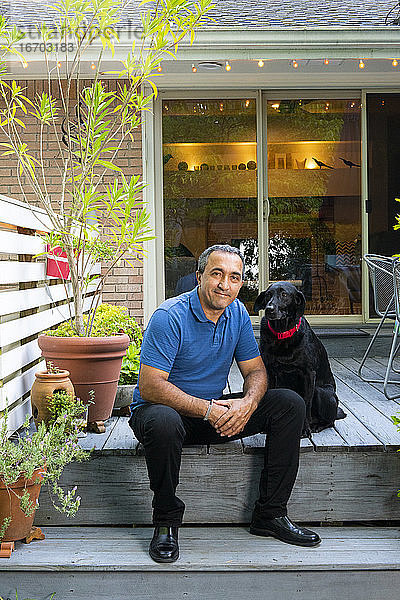 Lächelnder hispanischer Mann sitzt mit schwarzem Hund auf seiner Terrasse