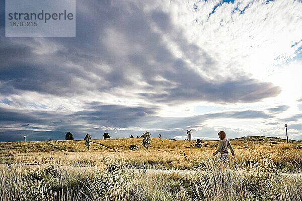 junges Mädchen geht durch ein Feld mit wildem Gras und einem dramatischen Himmel