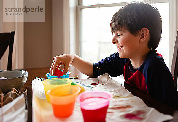 Ein fröhlicher Junge taucht ein Ei in eine Schale mit Farbstoff  um es für Ostern zu färben.