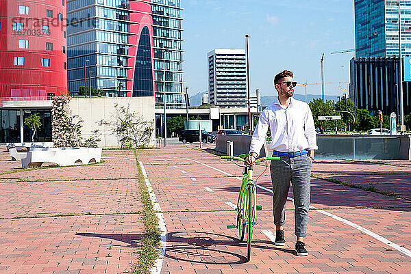 Bärtiger Mann mit Sonnenbrille  der mit seinem Fahrrad auf einem Fahrradweg im Freien spazieren geht