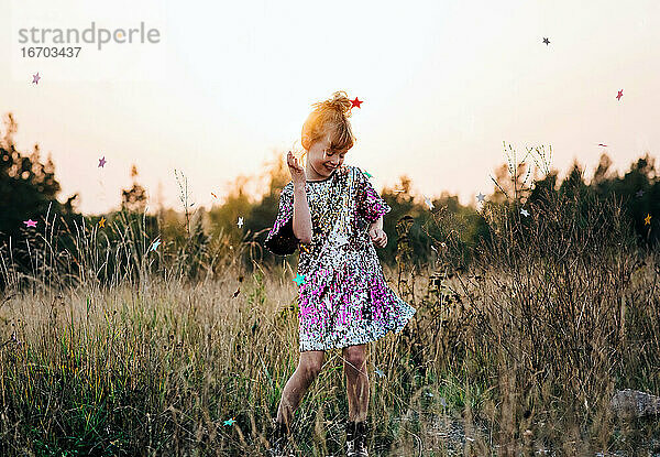 Mädchen tanzt fröhlich in einem glitzernden Kleid bei Sonnenuntergang mit Sternenkonfetti