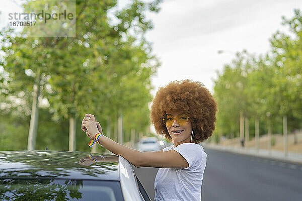 Frau mit Afro-Haar  die ein Foto mit ihrem Smartphone neben ihrem weißen Auto macht