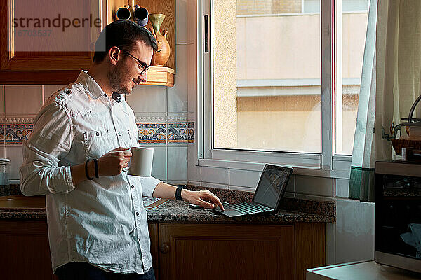 Junger Mann schaut auf seinen Laptop  während er in seiner Küche Kaffee trinkt