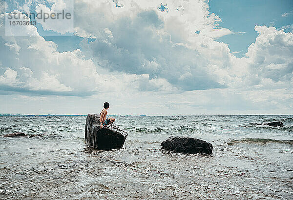 Ein Junge sitzt an einem heißen Sommertag auf einem großen Felsen in einem See.
