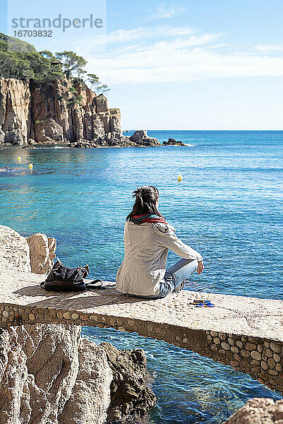 Rückansicht einer Frau  die sich vor dem blauen Meer entspannt  auf einer Brücke sitzend