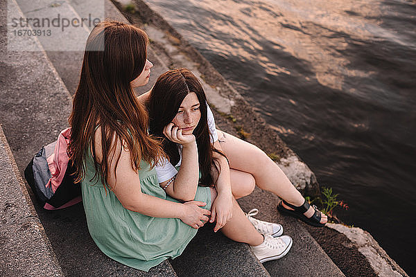 Freundinnen umarmen  während sie auf Stufen am Fluss in der Stadt sitzen