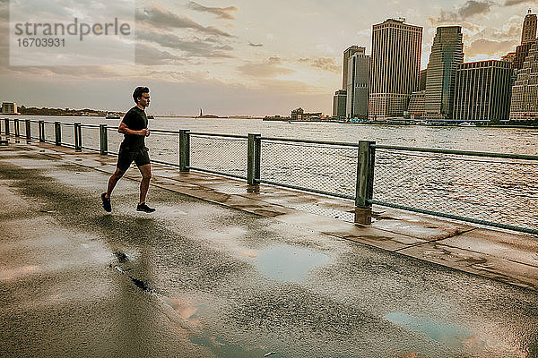 Männlicher Sportler  der bei Sonnenuntergang am Wasser läuft.