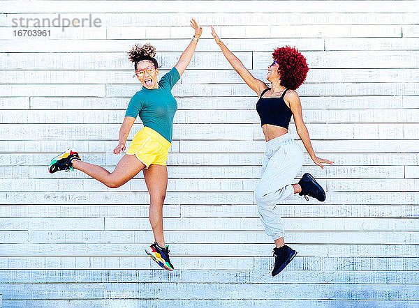 Zwei lateinamerikanische Frauen mit Afrofrisur  die auf einer weißen Wand springen und High Five machen.