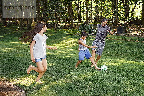 Ein Vater spielt mit seinen beiden Kindern barfuß im Garten Fußball