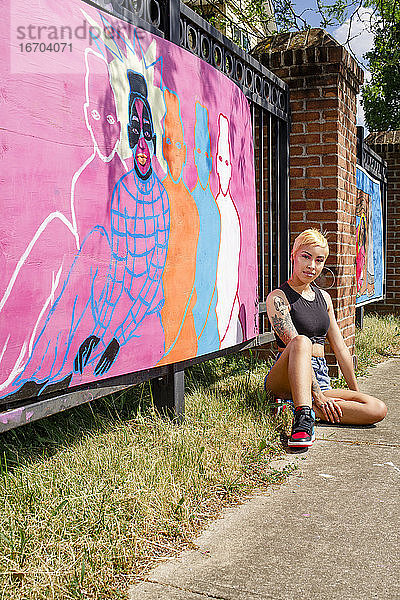 Eine stark tätowierte Künstlerin sitzt auf der Straße vor ihrem gemalten Wandbild