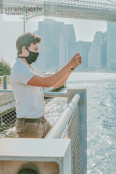 Junger Mann am Fluss mit Gesichtsmaske und Telefon.