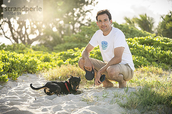 Junger fitter Mann hockt am Strand mit seinem kleinen schwarzen Hund in Hawaii