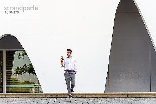 Junger bärtiger Mann lehnt an einer weißen Wand und benutzt ein Mobiltelefon