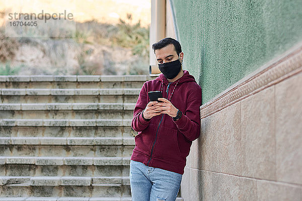 Junger Mann mit Maske schreibt auf seinem Handy und lehnt an einer Wand