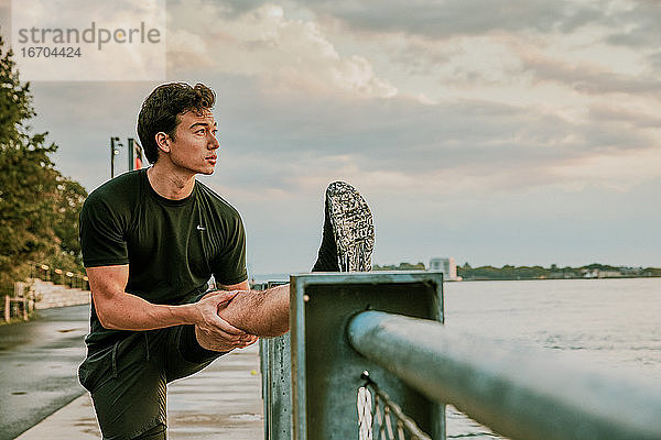 Männlicher Sportler  der sich bei Sonnenuntergang am Ufer dehnt.