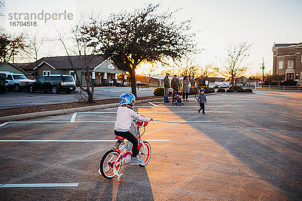 Junges Mädchen auf dem Fahrrad in Richtung Familie auf einem Parkplatz bei Sonnenuntergang