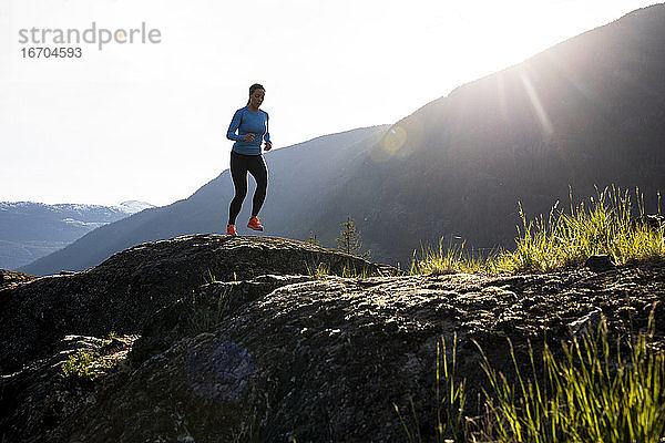 Vorderansicht einer Sportlerin in Sportkleidung  die an einem sonnigen Abend auf dem Lande auf Felsen in der Nähe von Bergen läuft