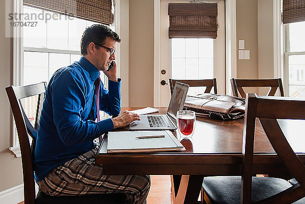 Mann am Handy  der in Pyjamahose von zu Hause aus an einem Computer arbeitet.