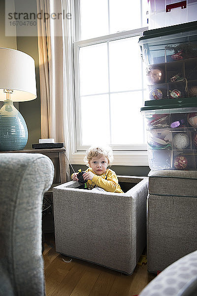 Kamerabewusstes Kleinkind sitzt in der Spielzeugkiste im Wohnzimmer vor dem Fenster