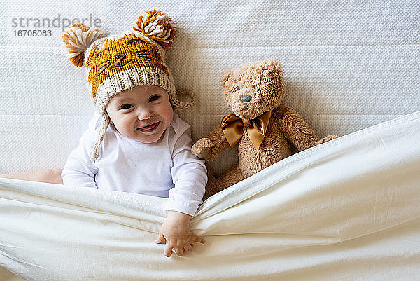 Glücklicher Junge mit Wollmütze liegt mit Teddybär unter der Decke auf dem Bett