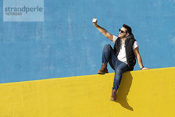 Junger bärtiger Mann mit Sonnenbrille sitzt an einer gelben Wand und macht ein Selfie