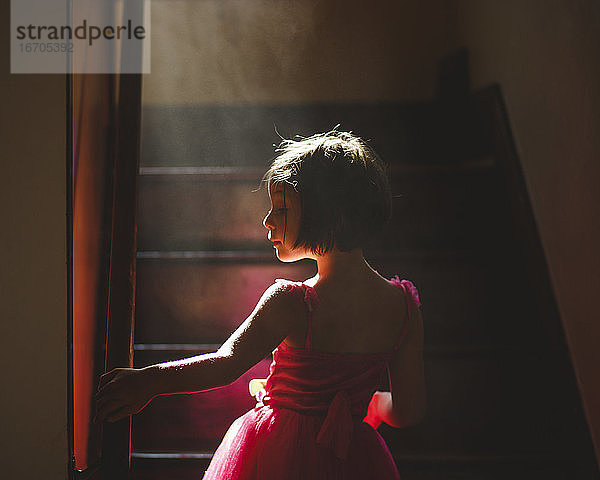 Ein anmutiges Kind steht in dunstigem Licht auf einer dunklen Treppe in einem ausgefallenen Kleid