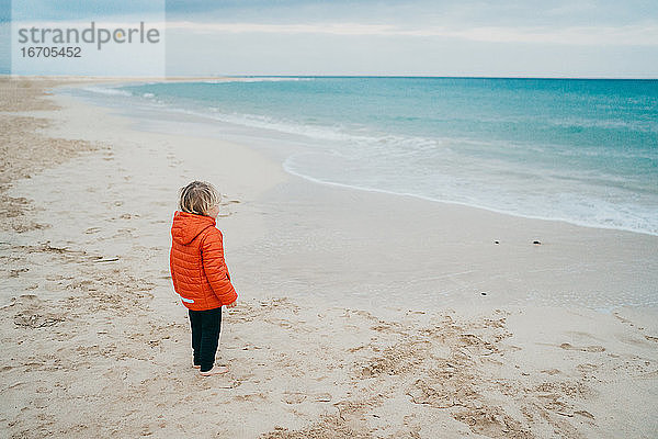 kleiner Junge mit Jacke am Strand an einem kalten Tag