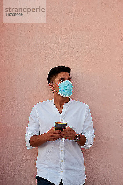 Junger lateinamerikanischer Mann mit Maske schaut auf sein Telefon auf rosa Hintergrund