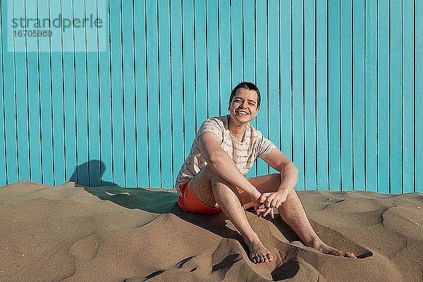 Junger Mann sitzt am Sandstrand und lächelt in die Kamera in einer türkisfarbenen Wand