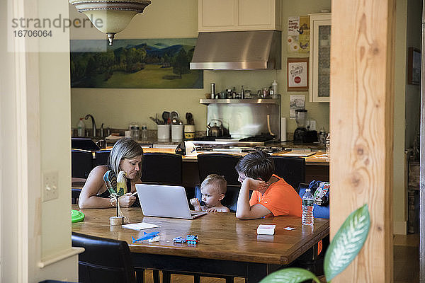 Wide View of Mom am Tisch mit Kleinkind Sohn auf Laptop und Teen Cousin