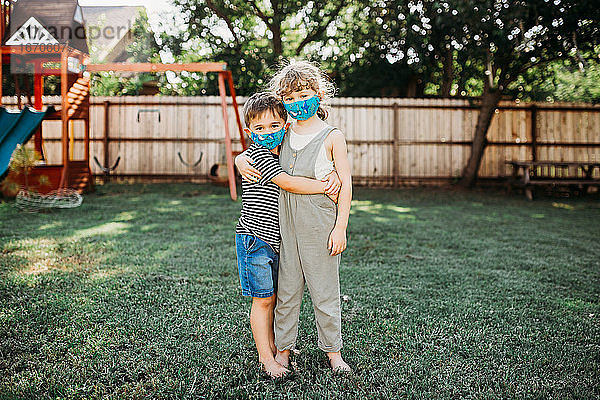 Zwei kleine Kinder umarmen sich draußen und tragen dabei selbstgemachte Stoffmasken