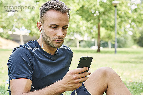 Mann mit seinem Smartphone  der nach dem Training in einem Park auf dem Rasen sitzt. Gesundes und gesundes Leben. Gesundheit und Sport Konzept.