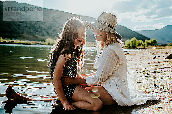 Mutter und Tochter lächelnd und kuschelnd am Seeufer an einem sonnigen Tag