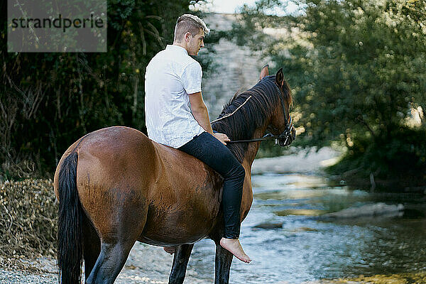 Porträt eines jungen blonden Mannes  der auf einem Pferd über einen Fluss reitet