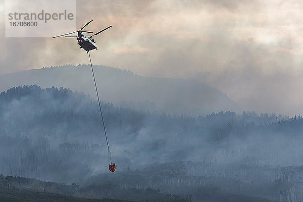 Militärhubschrauber fliegt mit Feuerschutzmittel  während Rauch von einem Waldbrand bei Sonnenuntergang aufsteigt