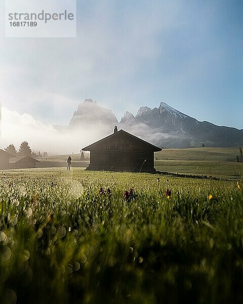 Frau seht auf nebliger Wiese neben Hütte  hinten Langkofel und Plattkofel  Morgenlicht  Seiser Alm  Südtirol  Italien  Europa