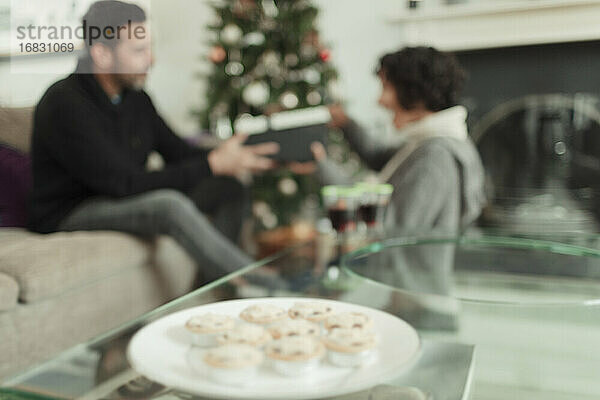 Ehefrau öffnet Weihnachtsgeschenk von Ehemann hinter Tablett mit Mince Pies